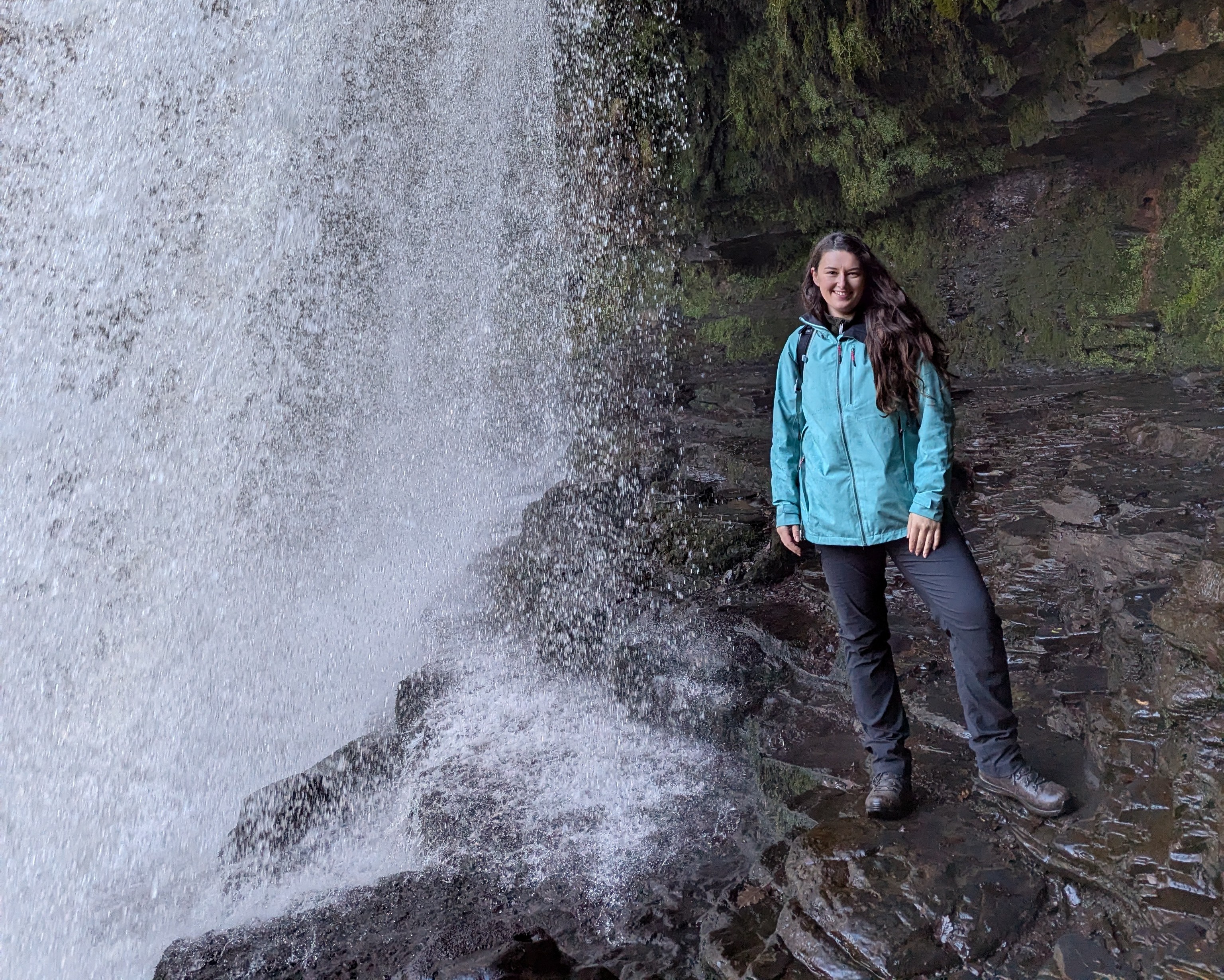 A woman with long dark brown hair, wearing a green waterproof jacket and black hiking trousers and brown hiking boots stands on a rock ledge behind a waterfall.